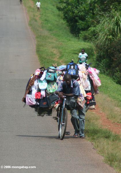 Verkäufer, der seine Waren auf einem Fahrrad trägt, das er drückt aufwärts entlang eine Landstraße in Uganda